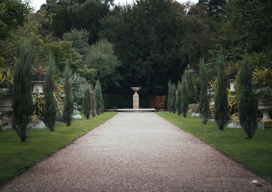A symmetrical garden pathway with a gravel surface running straight through well-maintained lawns on either side, bordered by young, narrow cypress trees. The path leads towards a classical stone pede