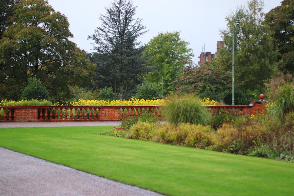 A well-maintained garden featuring a lush, neatly mowed green lawn in the foreground, bordered by a gravel pathway. Behind the lawn, there is a decorative red brick wall with a classic balustrade, top